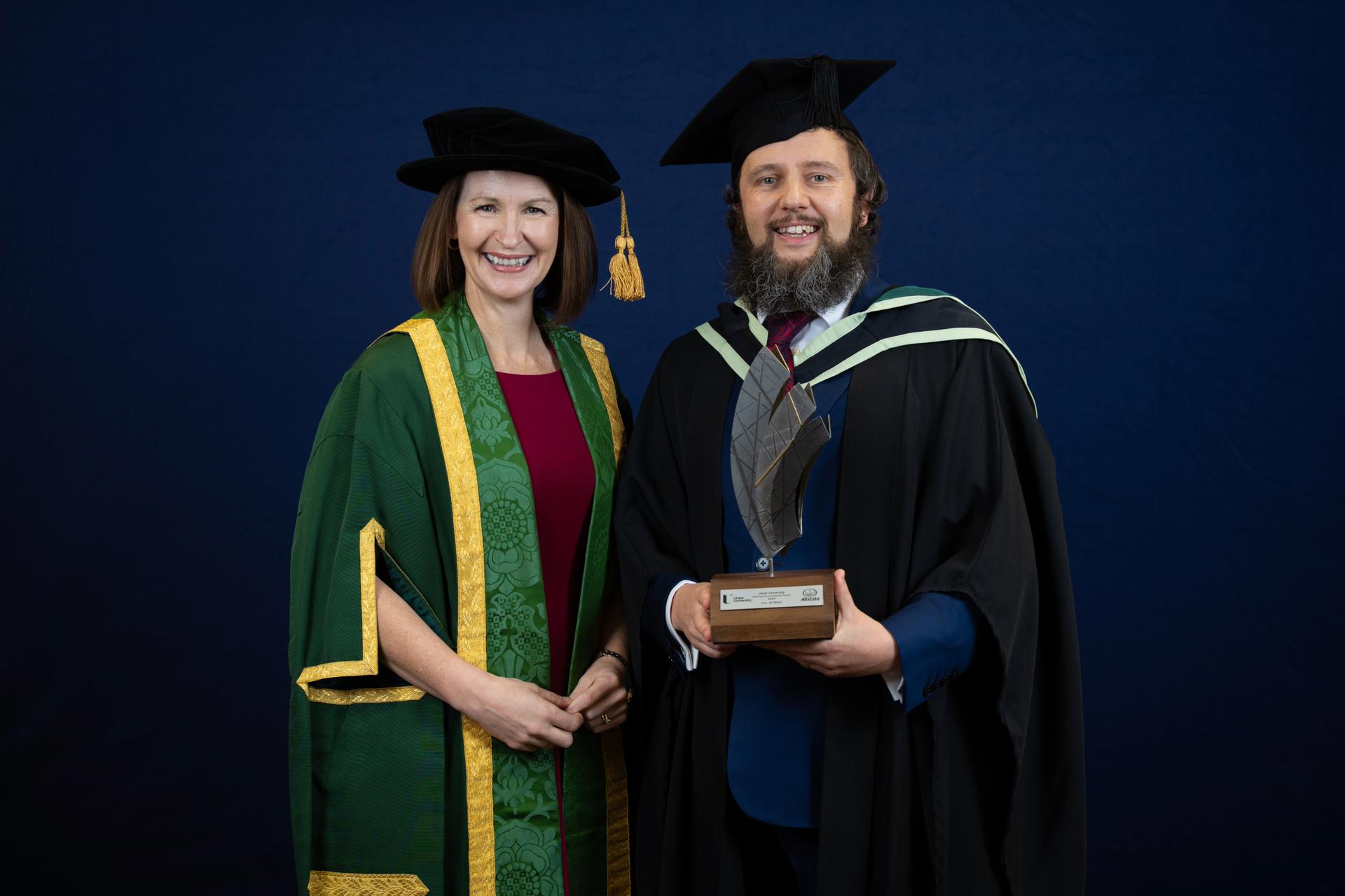 A smiling man and woman in academic gowns and caps stand against a navy background. The man holds an award, conveying a tone of achievement and celebration.